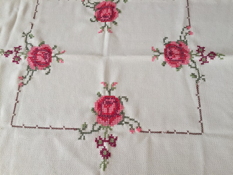 Puede incluir: Mantel blanco con un borde bordado marr&oacute;n y cuatro rosas rosas con hojas verdes.