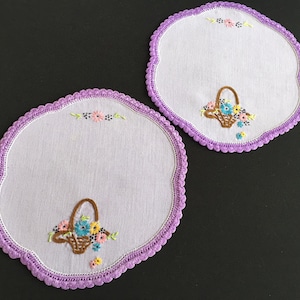 May include: Two white, round embroidered doilies with a scalloped purple border. Each doily features a brown basket filled with colourful flowers. Small embroidered flowers are also placed along the top edge.