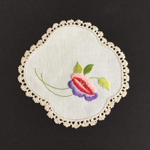 Pode incluir: Uma toalha de mesa branca, de formato quadrado, com borda de crochê. A toalha de mesa apresenta uma flor bordada com pétalas rosa, vermelhas e roxas, folhas verdes e caules verdes delicados. A borda de crochê é creme.
