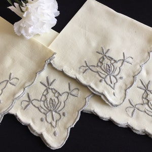 Puede incluir: Juego de tres servilletas de lino de color crema con bordes festoneados y diseños florales bordados en hilo plateado.