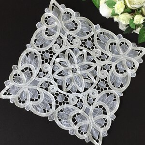 Puede incluir: Un tapete cuadrado de encaje blanco con un intrincado diseño calado, un motivo floral central rodeado de patrones de hojas en espiral y aberturas geométricas. La delicada tela tiene bordes cosidos y un aspecto semitransparente.