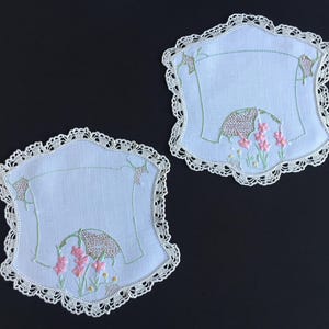 Puede incluir: Dos posavasos bordados en azul claro con un borde de encaje festoneado. Cada posavasos presenta un diseño floral con flores rosas, una cesta tejida y tallos verdes. Los posavasos están sobre un fondo negro.