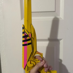 May include: A yellow toy pistol featuring a long barrel with black shading at the tip. The side panel displays a vibrant pink vertical stripe accented by horizontal black stripes. The handle and trigger guard are yellow with small pink striped details.