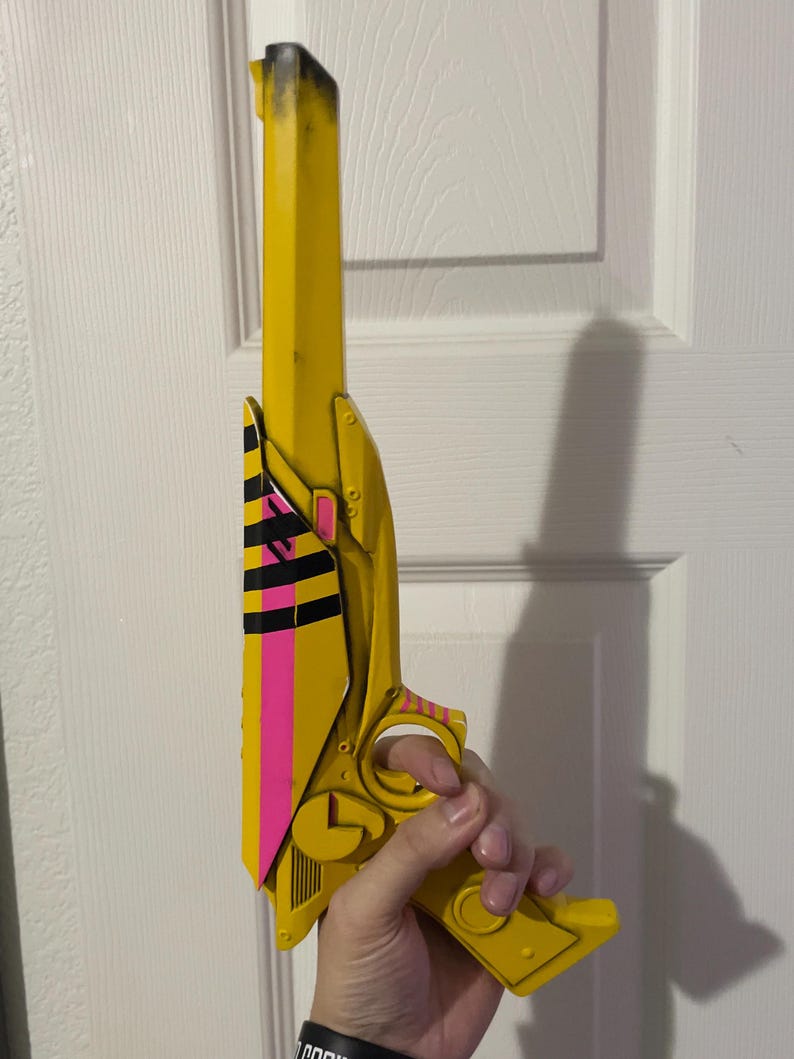 May include: A yellow toy pistol featuring a long barrel with black shading at the tip. The side panel displays a vibrant pink vertical stripe accented by horizontal black stripes. The handle and trigger guard are yellow with small pink striped details.