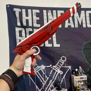 May include: A red and white prop blaster with a long barrel, a white handle, and a circular trigger guard. The blaster features a sleek, angular design with black weathering details along the edges and a striped pattern at the tip of the barrel.