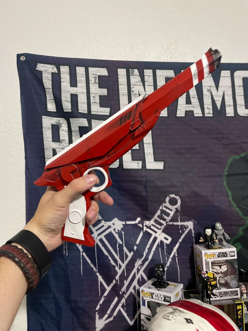 May include: A red and white prop blaster with a long barrel, a white handle, and a circular trigger guard. The blaster features a sleek, angular design with black weathering details along the edges and a striped pattern at the tip of the barrel.
