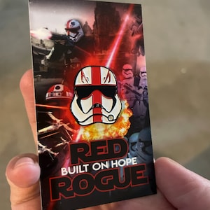 May include: A red and white enamel pin featuring a Star Wars Stormtrooper helmet with a red stripe down the middle. The pin is on a card with the text "Red Built on Hope Rogue".