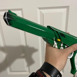 May include: A green prop pistol featuring a long barrel with black weathered detailing, white accents, and a small yellow rectangular element near the frame. The design includes a white grip section and a distinct muzzle shape with a triangular opening.