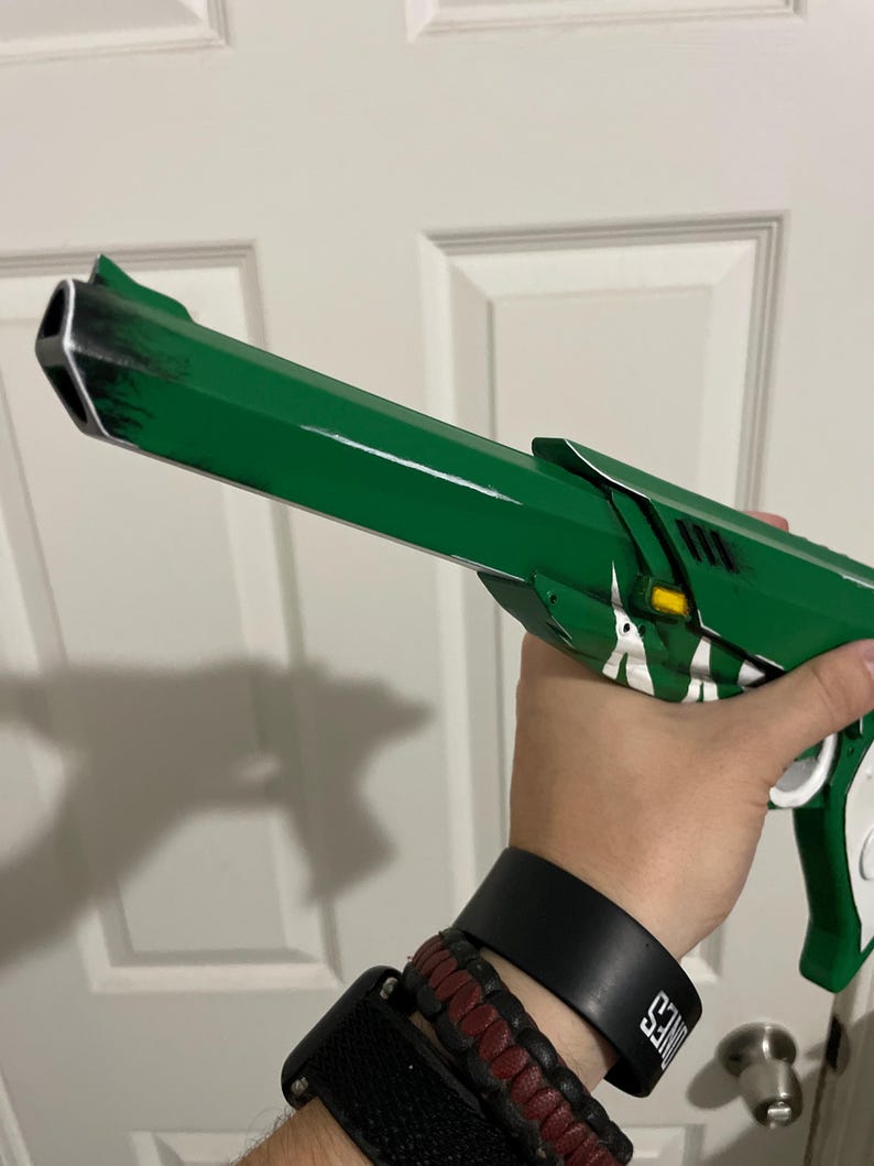 May include: A green prop pistol featuring a long barrel with black weathered detailing, white accents, and a small yellow rectangular element near the frame. The design includes a white grip section and a distinct muzzle shape with a triangular opening.