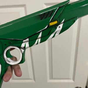 May include: A green prop pistol featuring a white circular trigger guard, a yellow rectangular accent, and black vent details. The body of the firearm is painted in a vibrant green finish with white decorative markings along the side paneling.
