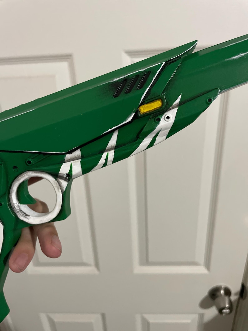 May include: A green prop pistol featuring a white circular trigger guard, a yellow rectangular accent, and black vent details. The body of the firearm is painted in a vibrant green finish with white decorative markings along the side paneling.