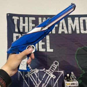 May include: A blue and white toy blaster with a long barrel, a white grip, and a trigger mechanism. The blaster features a sleek, angular design with a white stripe running along the top of the barrel and a circular white detail on the handle.