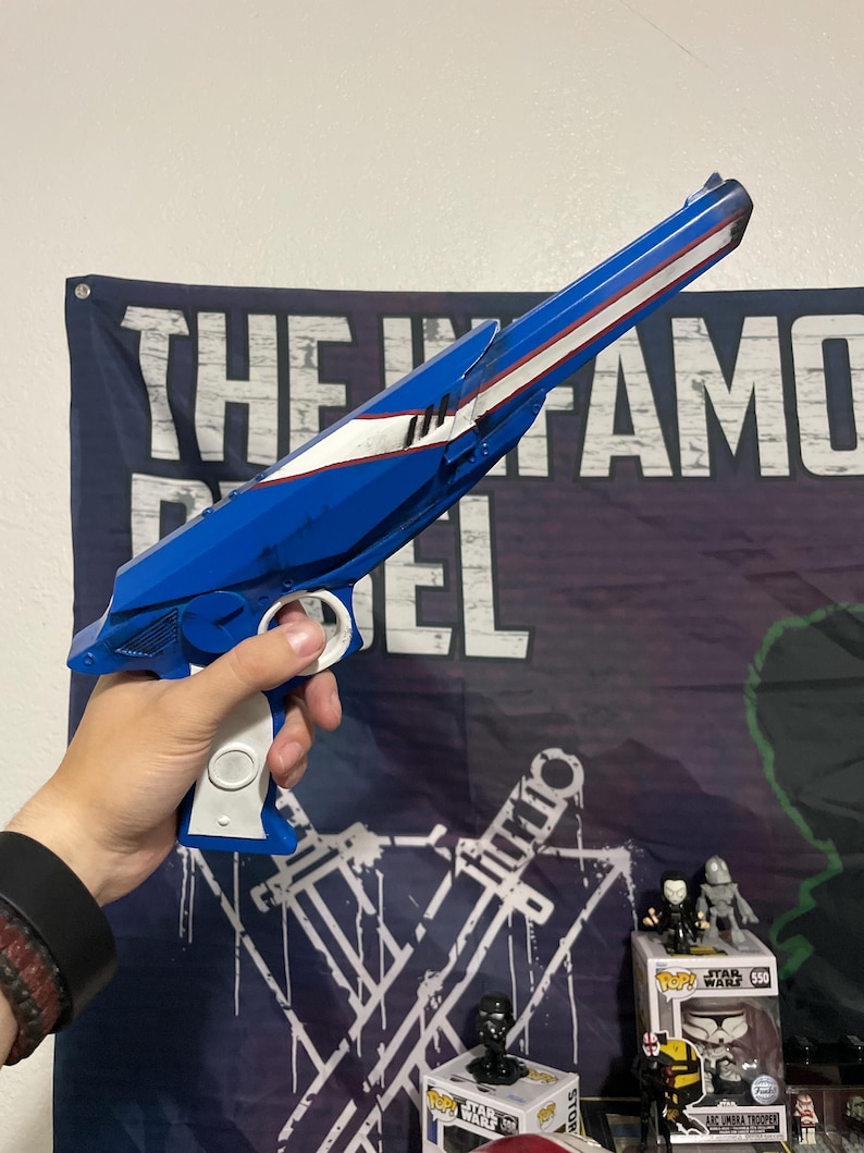May include: A blue and white toy blaster with a long barrel, a white grip, and a trigger mechanism. The blaster features a sleek, angular design with a white stripe running along the top of the barrel and a circular white detail on the handle.