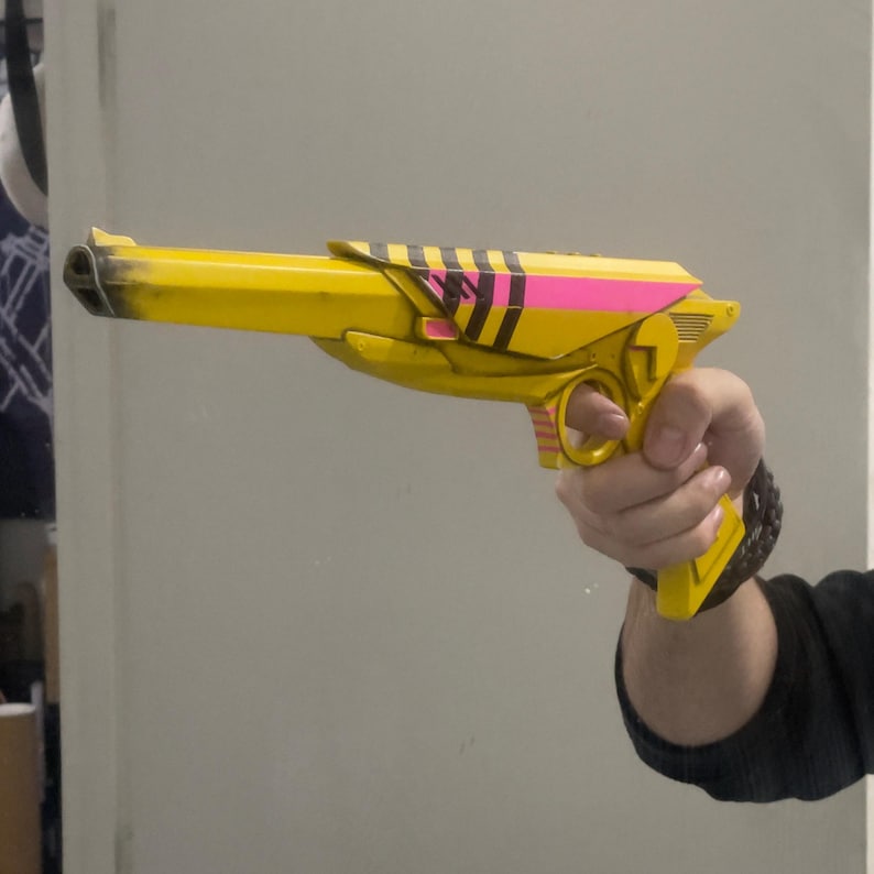 May include: A bright yellow toy pistol featuring a long barrel with black weathering at the tip. The body includes a vibrant pink horizontal stripe, black diagonal hazard stripes, and a trigger guard. The design has a futuristic aesthetic.