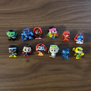 May include: A collection of eleven small, stylised superhero and villain figurines. The figures feature diverse characters with distinct costumes, including a green creature, a blue armoured robot, a red-hooded figure, and various masked heroes.