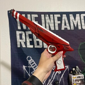 May include: A red and white toy blaster with a long barrel, a white trigger guard, and a white handle grip. The blaster features a sleek, angular design with a pointed muzzle and a distinct red and white striped pattern at the very tip of the barrel.