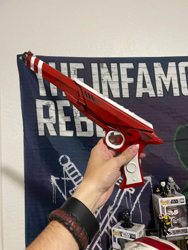 May include: A red and white toy blaster with a long barrel, a white trigger guard, and a white handle grip. The blaster features a sleek, angular design with a pointed muzzle and a distinct red and white striped pattern at the very tip of the barrel.