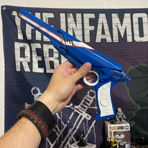 May include: A blue and white prop blaster with a long barrel, a white stripe running along the top, and a white handle grip. The blaster features a circular trigger guard and a mechanical design with angular edges, resembling a science fiction weapon.