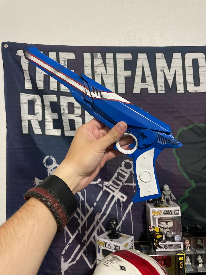 May include: A blue and white prop blaster with a long barrel, a white stripe running along the top, and a white handle grip. The blaster features a circular trigger guard and a mechanical design with angular edges, resembling a science fiction weapon.