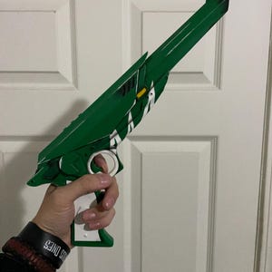 May include: A green and white toy blaster with a long barrel, a white trigger guard, and a white handle. The blaster features angular green panels, a small yellow rectangular accent, and white diagonal stripe detailing along the side of the main body.