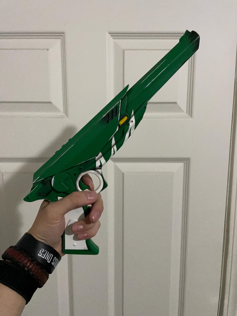 May include: A green and white toy blaster with a long barrel, a white trigger guard, and a white handle. The blaster features angular green panels, a small yellow rectangular accent, and white diagonal stripe detailing along the side of the main body.
