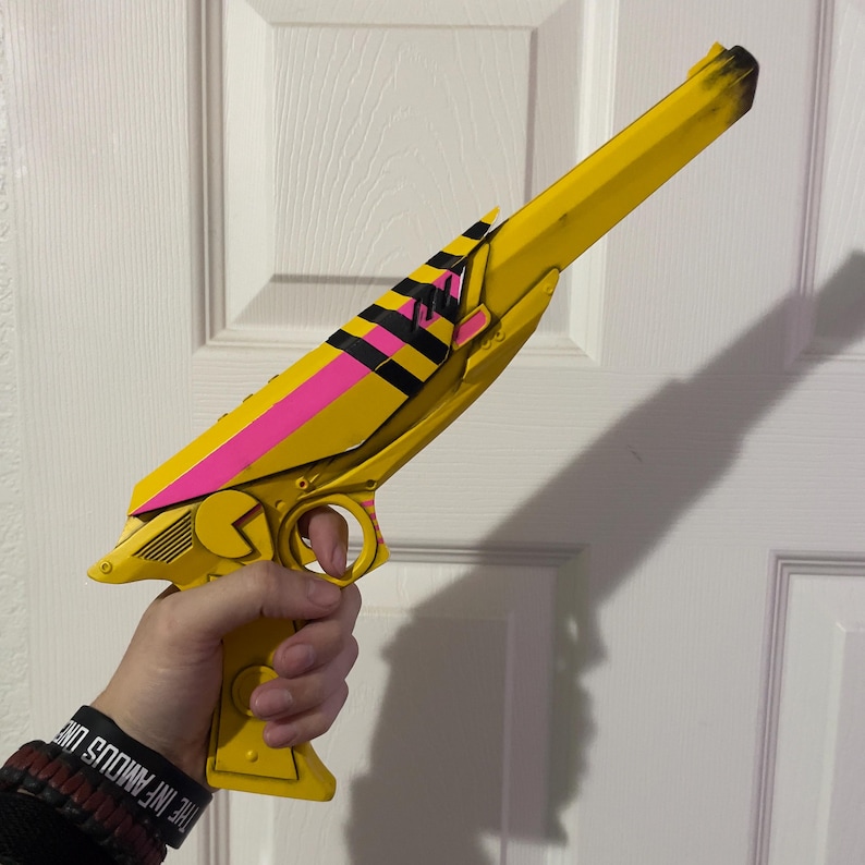 May include: A yellow toy pistol featuring a long barrel with black shading at the tip, a bright pink stripe along the top, and black and pink diagonal stripes on the central section. The handle includes a circular detail and a Pac-Man-like shape.