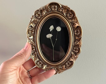 May include: An oval, antique-style gold frame with ornate detailing. Inside, against a black background, are delicate white flowers. The frame is being held up against a white wall.
