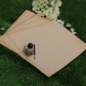 Op de afbeelding: Een stapel blanco, cr&egrave;mekleurige vellen papier met een klein glazen inktpotje en een paarse pen erop. Het papier ligt op een groene grasmat.