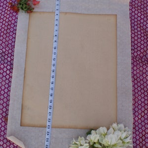 Op de afbeelding: Een wit stoffen frame met een beige papieren insert. Het frame ligt op een paars-witte gepatroneerde stof. Een witte meetlint is aan de rechterkant van het frame. Witte bloemen bevinden zich in de rechterbenedenhoek van de afbeelding.