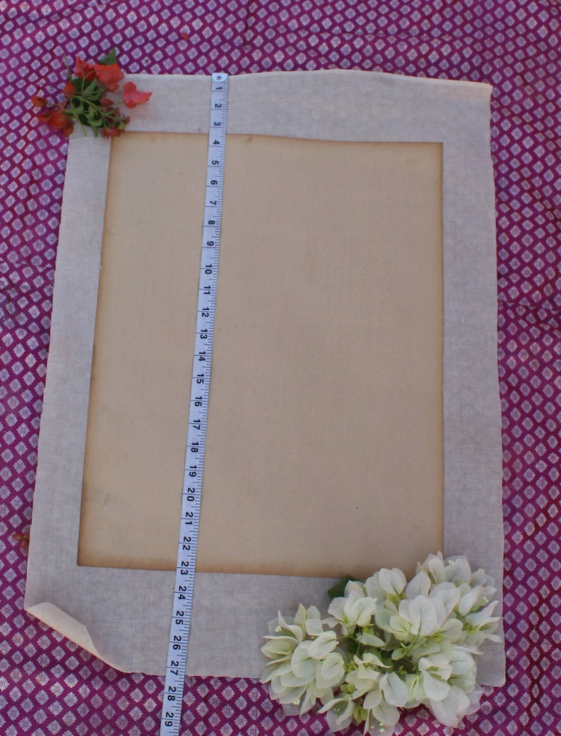 Op de afbeelding: Een wit stoffen frame met een beige papieren insert. Het frame ligt op een paars-witte gepatroneerde stof. Een witte meetlint is aan de rechterkant van het frame. Witte bloemen bevinden zich in de rechterbenedenhoek van de afbeelding.