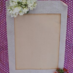 Op de afbeelding: Een wit stoffen frame met een bruine papieren insert. Het frame is versierd met witte bloemen in de linkerbovenhoek en rode bloemen in de rechterbenedenhoek. Het frame bevindt zich op een paarse en witte gepatroneerde achtergrond.