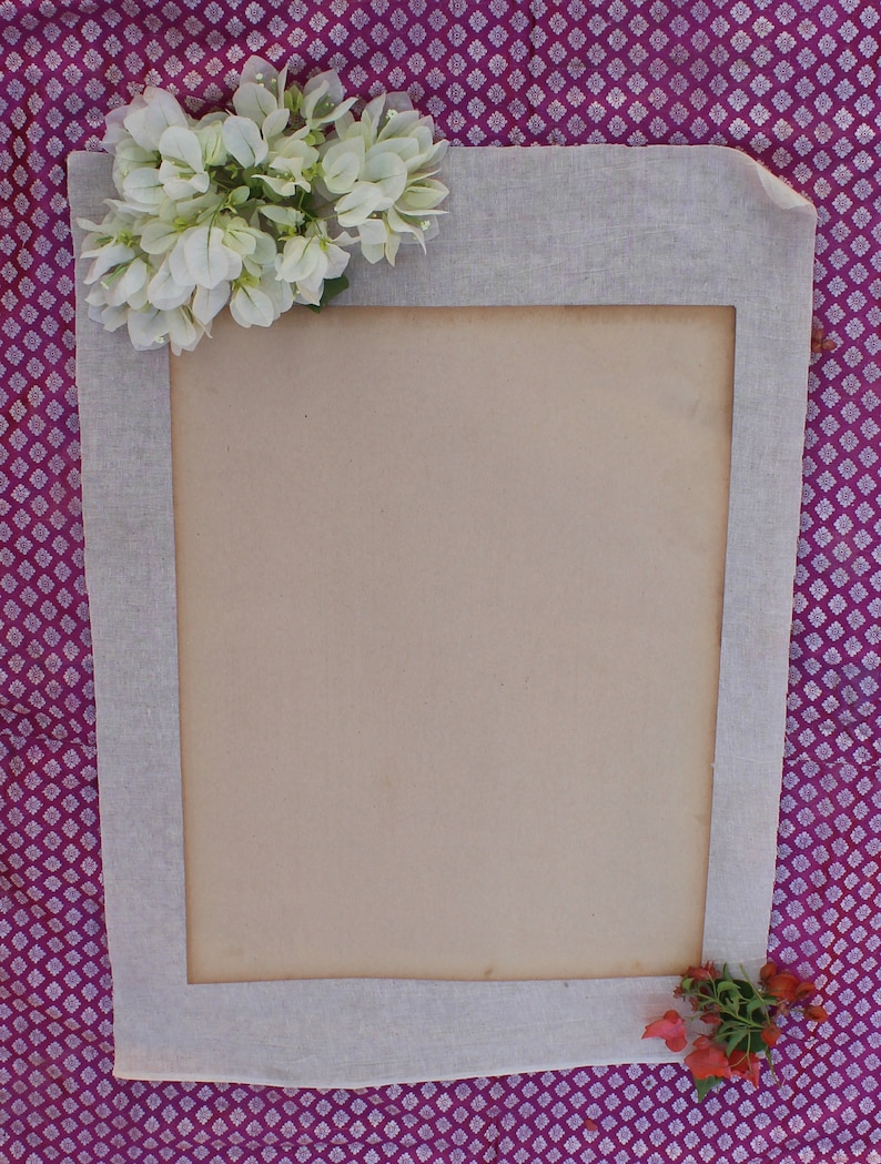 Op de afbeelding: Een wit stoffen frame met een bruine papieren insert. Het frame is versierd met witte bloemen in de linkerbovenhoek en rode bloemen in de rechterbenedenhoek. Het frame bevindt zich op een paarse en witte gepatroneerde achtergrond.