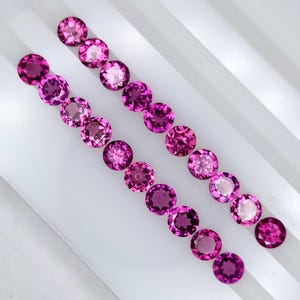 以下が含まれることがあります： 2列に並べられた16個の円形でファセットカットされた鮮やかなマゼンタピンクの宝石のコレクション。各石はブリリアントカットが施されており、複数のファセットが光を反射して、均一な円形の形状全体に輝きを生み出しています。