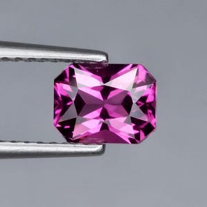May include: A vibrant, rectangular-cut magenta gemstone with a faceted surface that reflects light, held securely by a pair of silver-toned metal tweezers. The stone displays deep pink and purple hues with sharp, geometric edges and a polished finish.