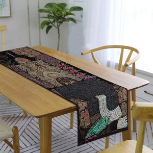 May include: A black table runner with a colourful, beaded design. The runner features a patchwork of different fabrics and embellishments, including sequins, beads, and embroidery. The runner is laid out on a wooden table.