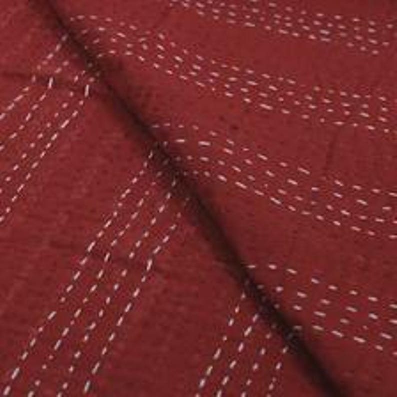 Puede incluir: Una tela de color rojo intenso con un patr&oacute;n de l&iacute;neas discontinuas blancas paralelas creadas con puntadas de hilv&aacute;n. El tejido tiene una textura visible y est&aacute; doblado sobre s&iacute; mismo, mostrando costuras lineales constantes.