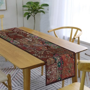 May include: A brown table runner with intricate embroidery and sequins. The runner has a patchwork design with various shades of brown, green, and gold. It is placed on a wooden table with a chair.