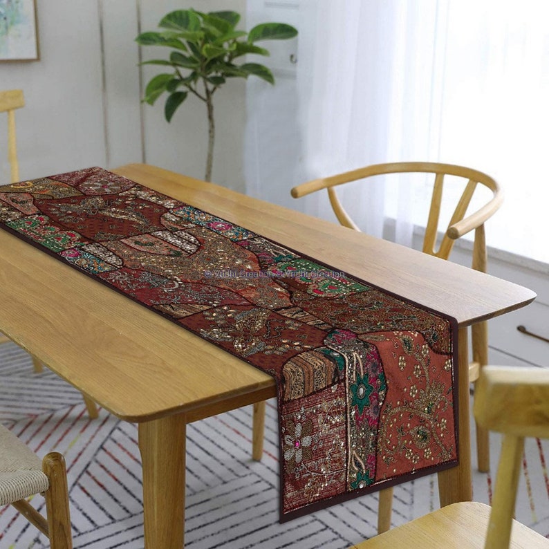 May include: A brown table runner with intricate embroidery and sequins. The runner has a patchwork design with various shades of brown, green, and gold. It is placed on a wooden table with a chair.