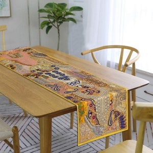 May include: A yellow table runner with a colourful embroidered design. The runner is made of fabric and has a beaded trim. It is placed on a wooden table with a chair.