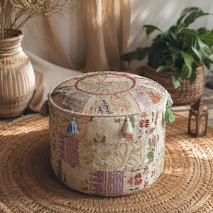 Puede incluir: Un puf de suelo redondo y bajo con un diseño de patchwork en telas de color crema, beige y estampados florales suaves. El taburete cilíndrico presenta costuras decorativas, un borde trenzado en la parte superior y pequeñas borlas colgantes en azul, blanco y verde.