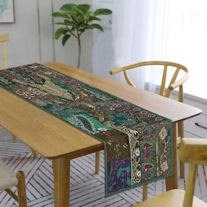 May include: A green table runner with intricate embroidery and sequins. The runner is made of patchwork fabric and features a variety of colours and patterns. It is displayed on a wooden table with a chair.