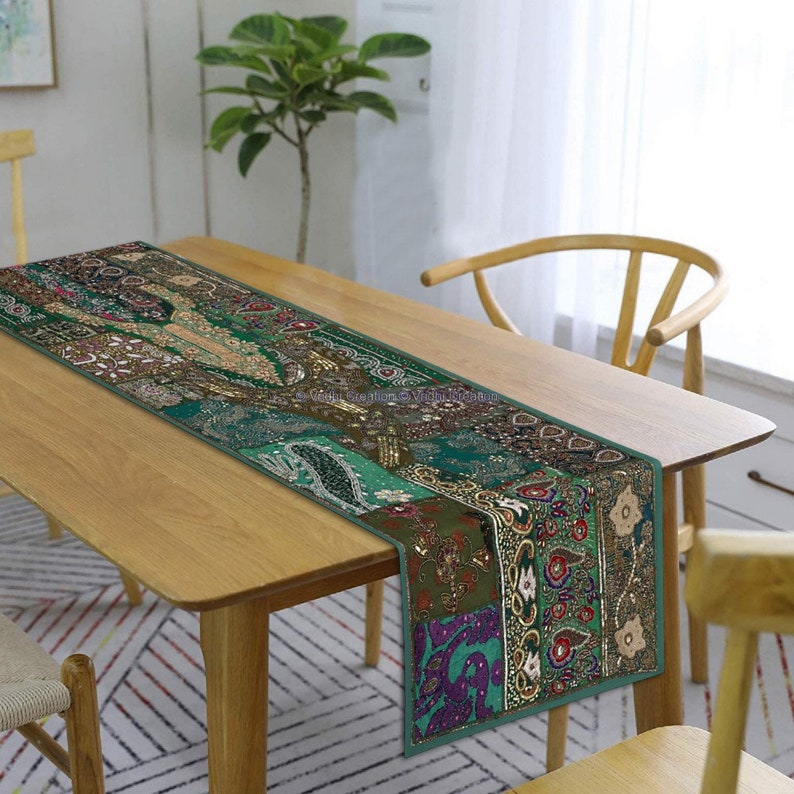 May include: A green table runner with intricate embroidery and sequins. The runner is made of patchwork fabric and features a variety of colours and patterns. It is displayed on a wooden table with a chair.