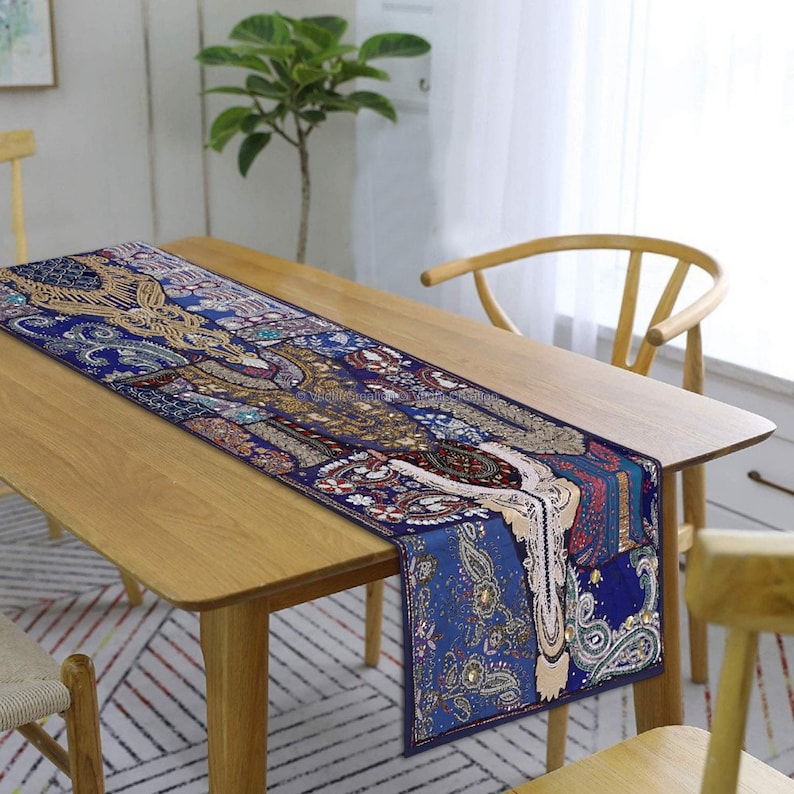 May include: A blue and gold table runner with a paisley pattern and beaded accents. The runner is on a wooden table with a chair.