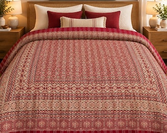 Op de afbeelding: Een rood en crème gekleurde quilt met een centraal blokprintontwerp en ingewikkelde geometrische en florale randen. De quilt ligt op een bed met twee grote crème kussens, twee donkerrode rechthoekige kussens en een klein lendenkussen.