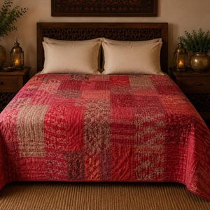May include: A patchwork quilt featuring a variety of red, burgundy, and muted tan fabric squares with intricate floral and geometric patterns. The quilt is stitched with a linear quilted texture and covers a bed with two plain beige pillows.