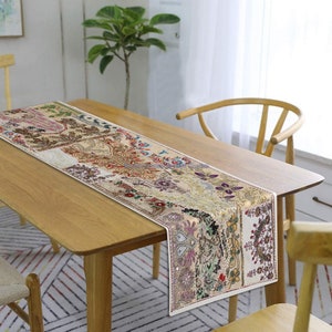 May include: A white table runner with a colourful patchwork design featuring floral and geometric patterns. The runner is made of fabric and has a subtle sheen. It is laid on a wooden table with a light wood finish.