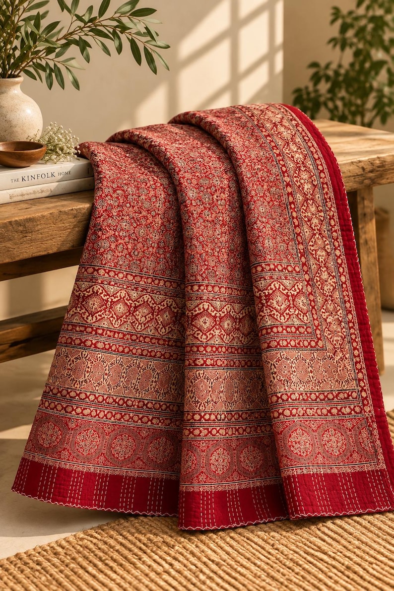 Puede incluir: Una manta acolchada de color rojo con intrincados patrones florales y geom&eacute;tricos en color crema. El tejido presenta un borde ancho con motivos circulares y detalles de costuras verticales en los extremos, creando un aspecto texturizado.