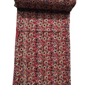May include: A rolled-up, red and beige floral patterned quilt. The quilt features a repeating pattern of beige flowers with black centers and beige leaves on a red background. The quilt is hand-stitched with white thread.