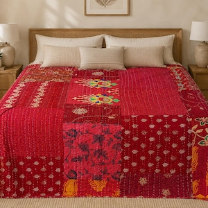 Op de afbeelding: Een patchwork sprei bestaande uit stoffen vierkanten in diverse tinten rood, roze en oranje. De sprei is voorzien van gedetailleerd kantha-stiksel en toont verschillende bloem-, blad- en geometrische patronen op de afzonderlijke stoffen delen.