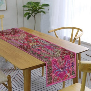 May include: A pink table runner with intricate embroidery and sequins. The runner is made of patchwork fabric and features a variety of colours and patterns. It is displayed on a wooden table with a light wood finish.